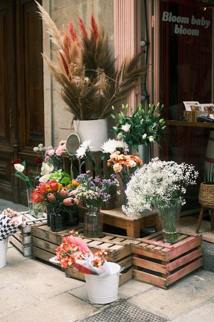 An outdoor display of fresh floral arrangements arranged on wooden pallets and in vases, featuring a variety of flowers including pink roses, white daisies, baby's breath, and mixed seasonal blooms in